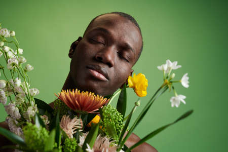 Young African man with closed eyes enjoying smell of flowers while holding bouquet in front of...
