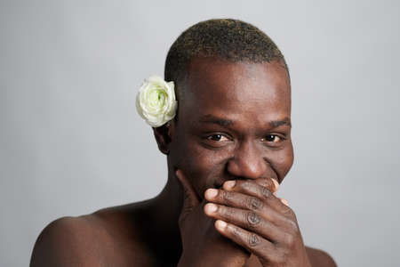 Young African man with white camelia flower by ear covering his mouth while laughing in front of camera in isolationの写真素材