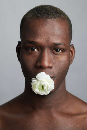 Young black man with bud of white camelia in his mouth standing in front of camera in isolation and...