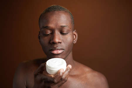 Young African man looking at moisturizing body cream in plastic jar while standing in front of camera over brown backgroundの写真素材