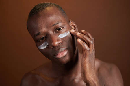 Young man of African ethnicity looking at you while applying moisturizing day cream on under-eye area in front of cameraの写真素材