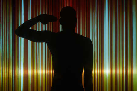 Black figure of young muscular shirtless man standing against striped wall in darkness and keeping...