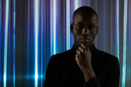 Serious young African man in black shirt touching his face by hand while standing against wall with...
