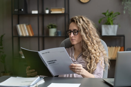 Young serious businesswoman looking through financial documents while sitting by desk against office environmentの写真素材