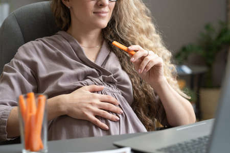 Contemporary pregnant businesswoman eating carrot and keeping hand on belly while sitting in front of laptopの写真素材