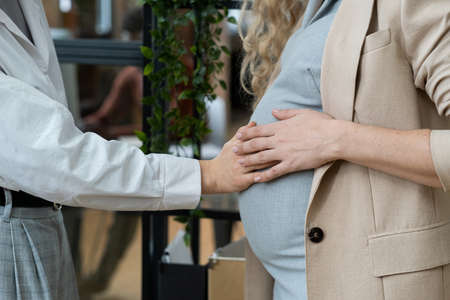 Young businesswoman touching belly of her pregnant colleague while both standing in front of camera in officeの写真素材
