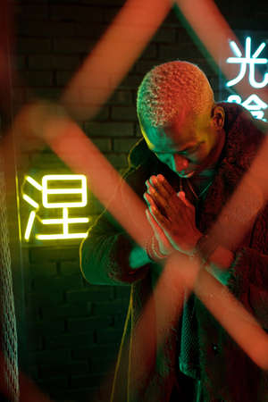 Young African man in furcoat keeping his hands put together and bowing while standing behind bars against illuminated hieroglyphsの写真素材