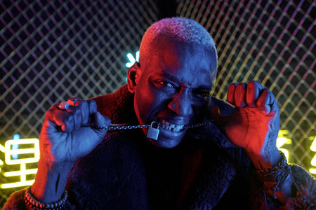 Young African man with chain between his clenched teeth standing in front of camera against bars and looking at youの写真素材