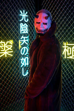 African man in monster mask and long brown furcoat looking at camera while standing against bars...