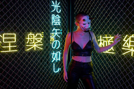 Young cyberpunk female in monster mask and black leather clothes holding by bars while standing in front of cameraの写真素材