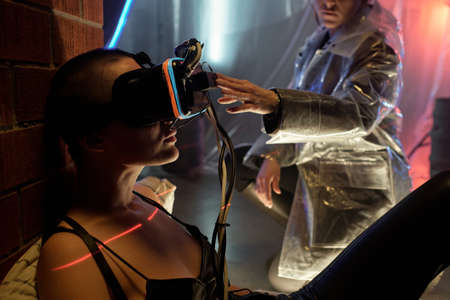 Hand of young woman touching virtual reality headset on head of cyberpunk girl in black leather...