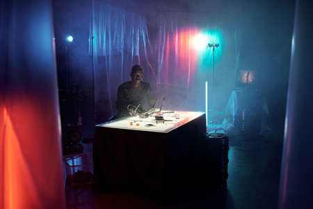 Young African man sitting by table with electronic equipment inside hacker hideouts illuminated with...