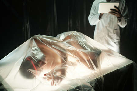 Young African male scientist or researcher with tablet standing by table with cyberpunk girl lying under cellophane in labの写真素材