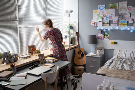 Sick teenage girl looking through the window while standing in her equipped hospital wardの写真素材