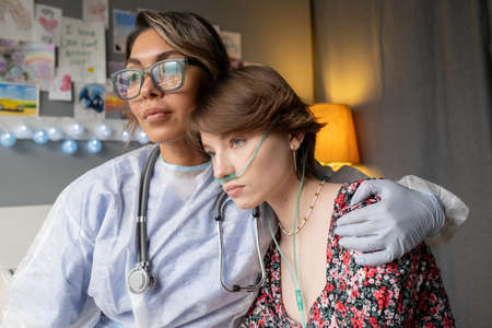 Young nurse in protective wear embracing the sad sick girl, she supporting her during disease at hospital wardの写真素材