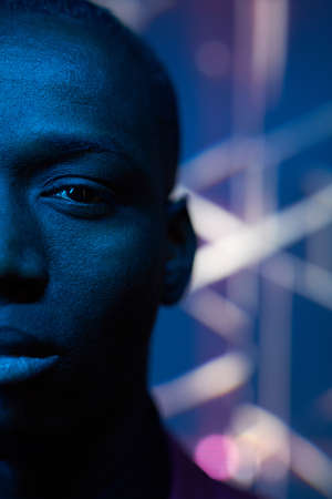 Close-up of African young man looking at camera with blank expression in dark studio with blue light