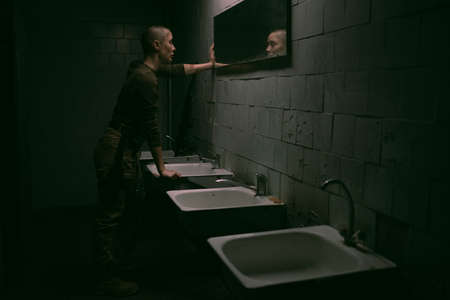 Wide angle side view at tough woman with buzzcut looking at mirror in dark survival setting, copy...