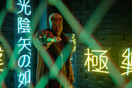 Young man standing behind bars and pointing with gun at camera against illuminated hieroglyph meaning nirvana and western paradise in darknessの写真素材