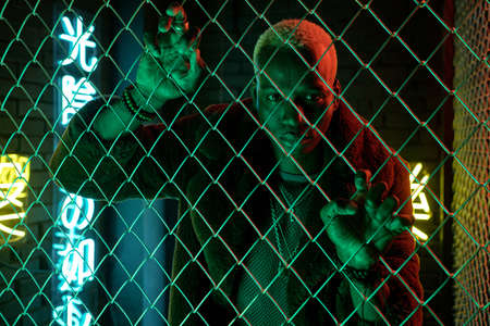 Portrait of African cyberpunk man looking at camera standing behind the bars in dark with neon...