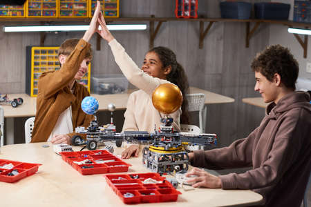 Group of teenage kids high fiving while enjoying building robots in engineering class at school,...