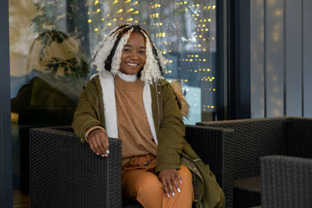 Portrait of African girl in warm clothing smiling at camera while resting on armchair on the porch...