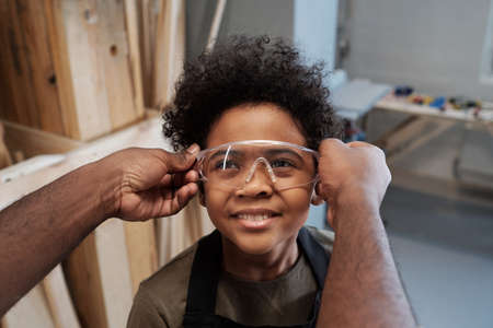 Close up of unrecognizable African-American man giving protective goggles to cute son helping father in workshopの写真素材