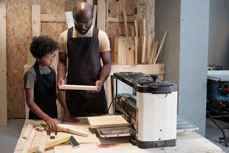 Waist up portrait of African-American father and son using electric saw machine while working...