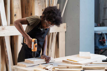 Waist up portrait of young black boy in carpentry workshop using power drill while building furniture, copy spaceの写真素材