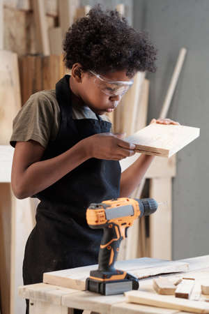 Vertical portrait of young black boy with curly hair inspecting piece of wood in carpentry workshopの写真素材