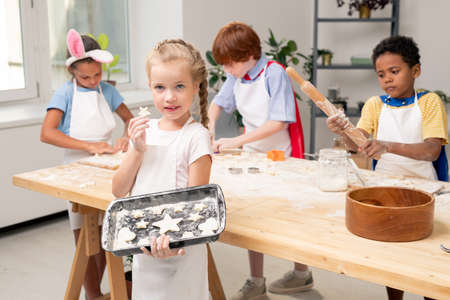 Cheerful curly little girl in apron crossing arms on chest while standing by kitchen table with ingredients for cookiesの写真素材