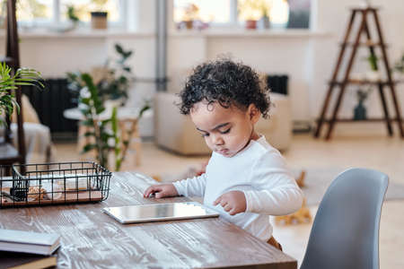 Concentrated modern mixed race toddler with curly hair standing at desk and using tablet in living roomの写真素材
