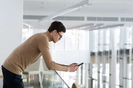Young serious employee in beige casual pullover bending over railing in office building while texting in smartphone or searching through contactsの写真素材