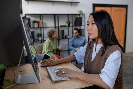 Young Asian businesswoman in casual clothes working in front of computer monitor on background of two intercultural coworkersの写真素材