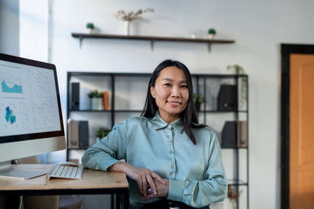 Young Asian female economist in smart casualwear standing by workplace with computer monitor and...