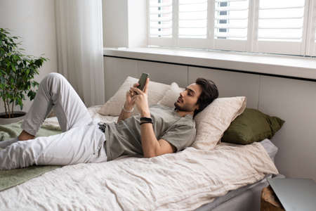 Contemporary young manager or businessman in casualwear texting in smartphone while lying on bed during work from homeの写真素材
