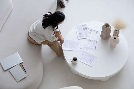 Overview of young brunette female accountant looking through financial papers while sitting on sofa and bending over white round tableの写真素材