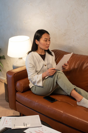 Young serious Asian female auditor looking through financial documents while sitting on brown...