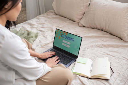 Young female student in casualwear sitting on bed and entering her name and password to log in for online course of studyの写真素材