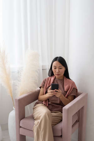 Young serious Asian woman in casualwear texting in mobile phone while sitting in armchair by window...