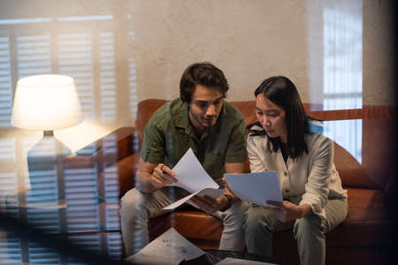 Young Asian woman with documents consulting with male coworker at meeting in home office while sitting on leather sofaの写真素材