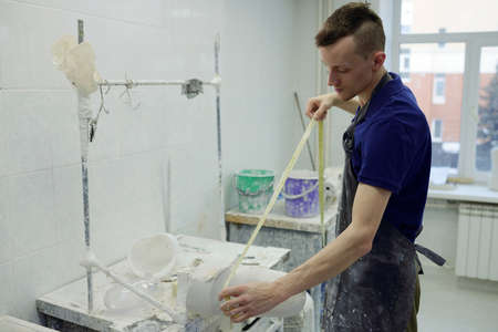 Young male worker in apron measuring plaster cast for leg prosthesis while standing by workplace in...