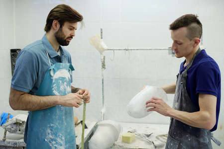 Young man in apron showing coworker prosthetic socket and consulting with him about size and fitting...