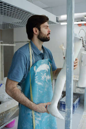 Serious young specialist of prosthetic factory holding plaster cast of leg and looking at it while standing by display with artificial limbsの写真素材