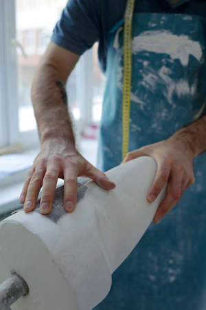 Hand of male specialist in apron rubbing surface of plaster workpiece of leg part with sandpaper in...