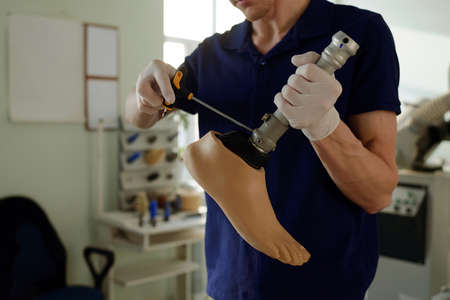 Young gloved manual worker of factory assembling parts of artificial leg and fixing steel shaft to...
