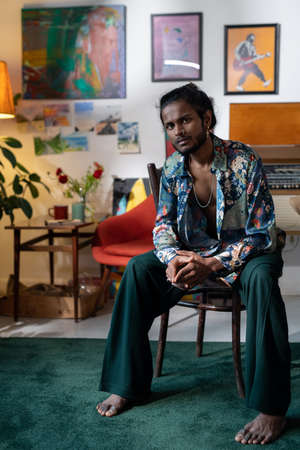 Young relaxed barefoot man in fashionable casualwear sitting on chair in modern art or design studio...