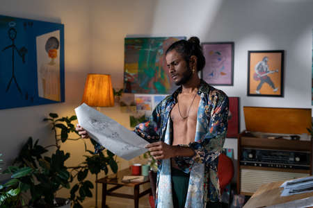 Young inspirated man in non-gendered clothing looking at drawing while standing in studio of modern...
