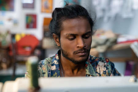 Young contemporary designer of clothes sitting in front of electric sewing machine and working over...