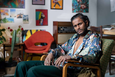 Young serious non-binary man in trendy clothes sitting in armchair in front of camera while having rest in modern art studioの写真素材