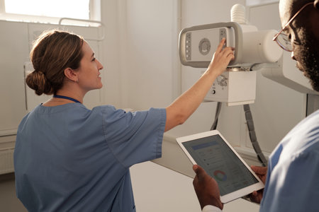 Young female clinician in uniform pushing button on panel of x-ray machine while preparing for examination of patients in hospitalの写真素材